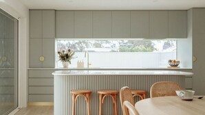 Una cocina moderna con armarios grises, una encimera blanca y una ventana con vistas a un patio trasero. Hay tres taburetes de mimbre y una mesa de comedor con una jarrón blanco.