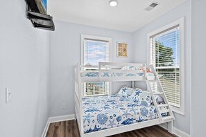 A bunk bed with a nautical-themed bedspread, two windows with blinds, and a wall-mounted shelf.