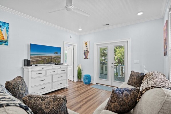 A living room with a white entertainment unit, a large flat-screen TV, a sofa with patterned cushions, and a sliding glass door leading to an outdoor area.