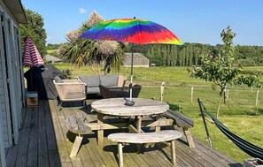 Une terrasse en bois avec une table ronde et des bancs, un parasol coloré et du mobilier d’extérieur.