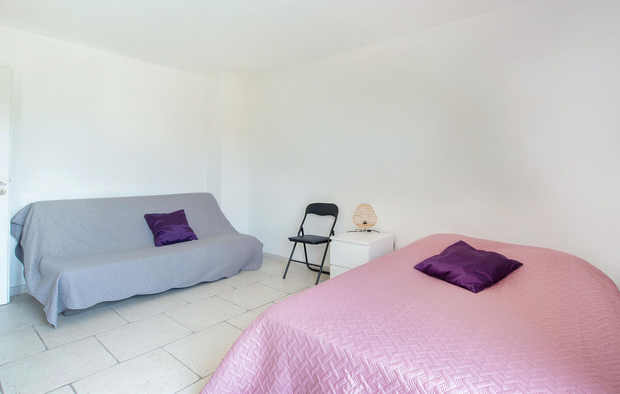 Une chambre avec deux lits, l’un recouvert d’une couvre-lit rose et l’autre d’une couvre-lit grise. On y trouve une chaise noire et une petite table blanche avec une lampe.