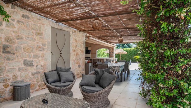 Un patio couvert, aménagé avec des meubles en osier, des murs en pierre et offrant une vue sur le jardin.