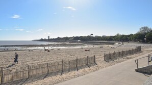 A beach with a wooden fence, a concrete walkway, and a distant view of buildings and trees. - Appartement Proche Plage (La Rochelle)