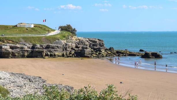 A coastal beach with a rocky cliff, a path leading to a building with a flag, and the sea in the background.