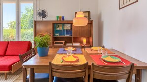 A dining area with a wooden table set for four, surrounded by wooden chairs. A red sofa is visible to the left, and a blue pot with flowers is on the table.