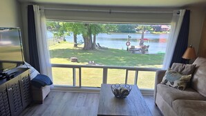 A living room with a sofa, a coffee table, and a TV on a cabinet. A window offers a view of a lake and outdoor furniture. - Charming 3-bedroom cottage in serene Trent Hills with WiFi (Trent Hills)