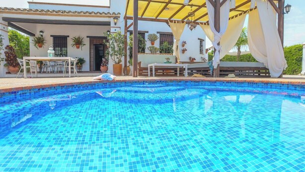 Ein klares, blaues Schwimmbecken mit Fliesenboden, umgeben von einer überdachten Terrasse mit Gartenmöbeln und Blumentöpfen.