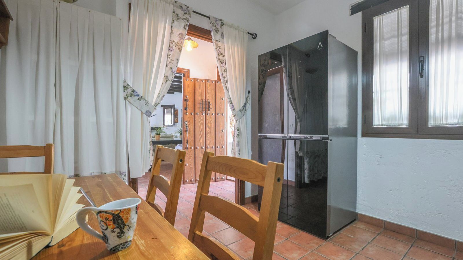 A kitchen with a wooden table, chairs, a refrigerator, and a window with curtains.