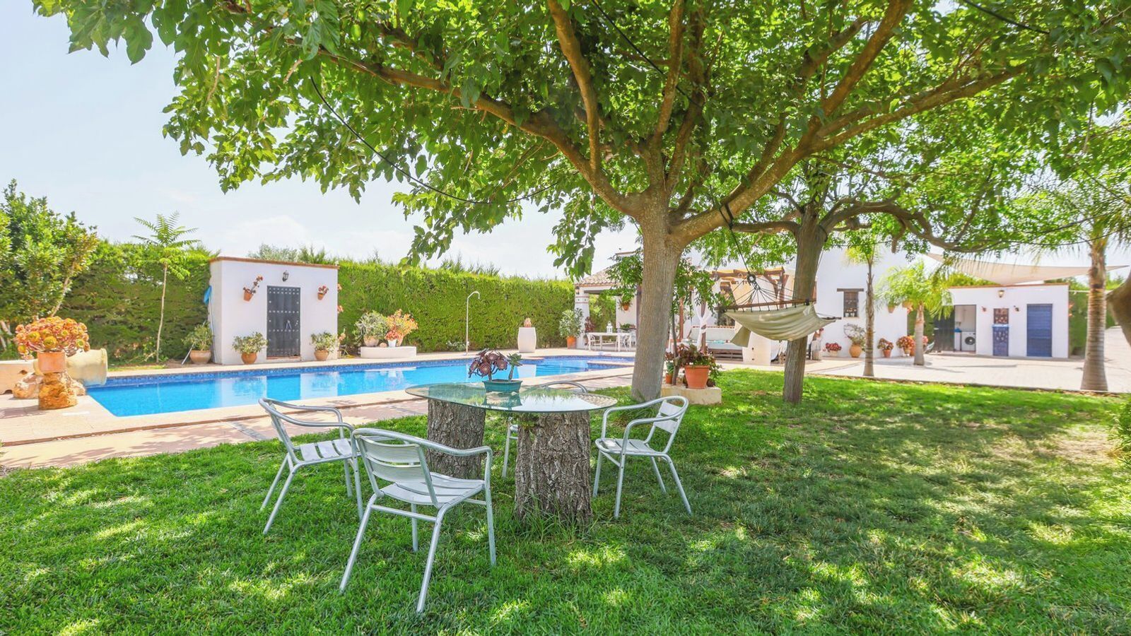 A pool area with a glass table and chairs, surrounded by well-maintained grass and trees.