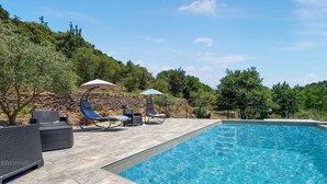 Une piscine entourée d’un mur de pierre et de verdure, avec des chaises longues et des parasols.