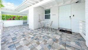 Un patio cubierto con una puerta blanca que dice "1910", una pequeña mesa con dos sillas, y vista a un seto verde.