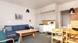 Een gezellige, compacte leefruimte met een bank, een eettafel met stoelen, een kitchenette en een schilderij aan de muur.