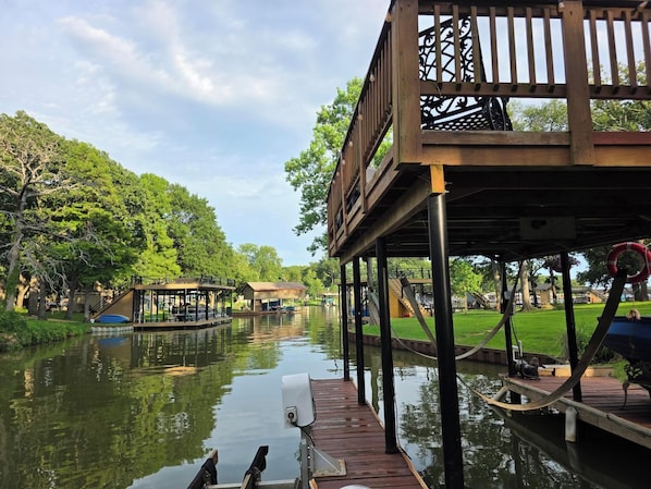 A wooden deck with a view of a calm river, surrounded by lush greenery and a clear sky.
