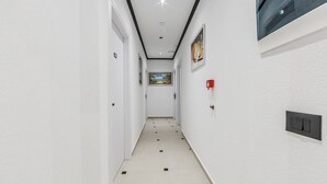 Un couloir étroit aux murs blancs, doté d’un sol carrelé et d’un luminaire au plafond. Des portes se trouvent de part et d’autre, un détecteur d’incendie est fixé au mur de droite, et un tableau encadré est placé au-dessus du détecteur d’incendie.