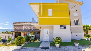 Une maison à deux étages avec une façade jaune et blanche, une porte d’entrée et un petit jardin agrémenté de plantes en pot.