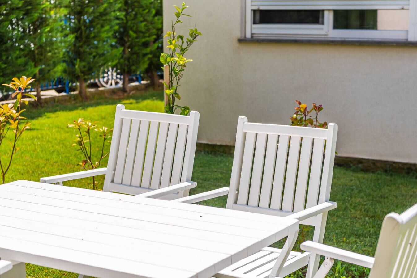 Een witte houten tafel en twee witte stoelen buiten, met een gazon en een gebouw op de achtergrond.