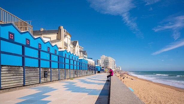 A coastal promenade with a blue sea wall, a sandy beach, and a clear blue sky.