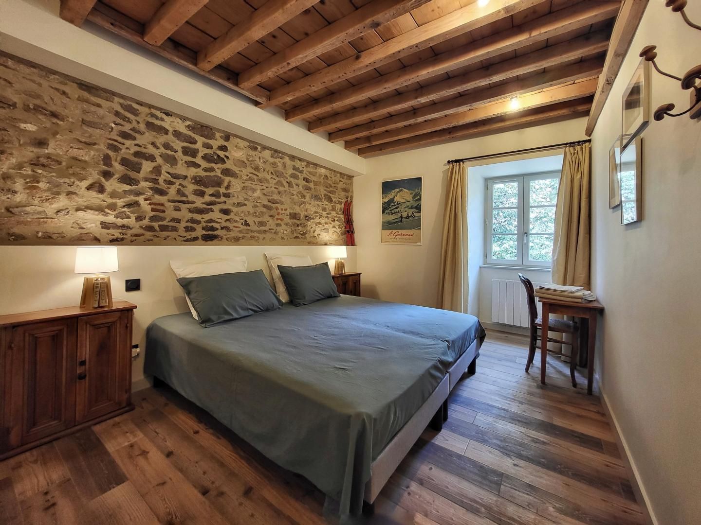 Ein Schlafzimmer mit Steinwand, Holzbalken, einem Bett mit Kissen, einem Nachttisch, einem Stuhl und einem Fenster mit Vorhängen.