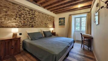 Un dormitorio con pared de piedra, vigas de madera, cama con almohadas, mesita de noche, una silla y ventana con cortinas.