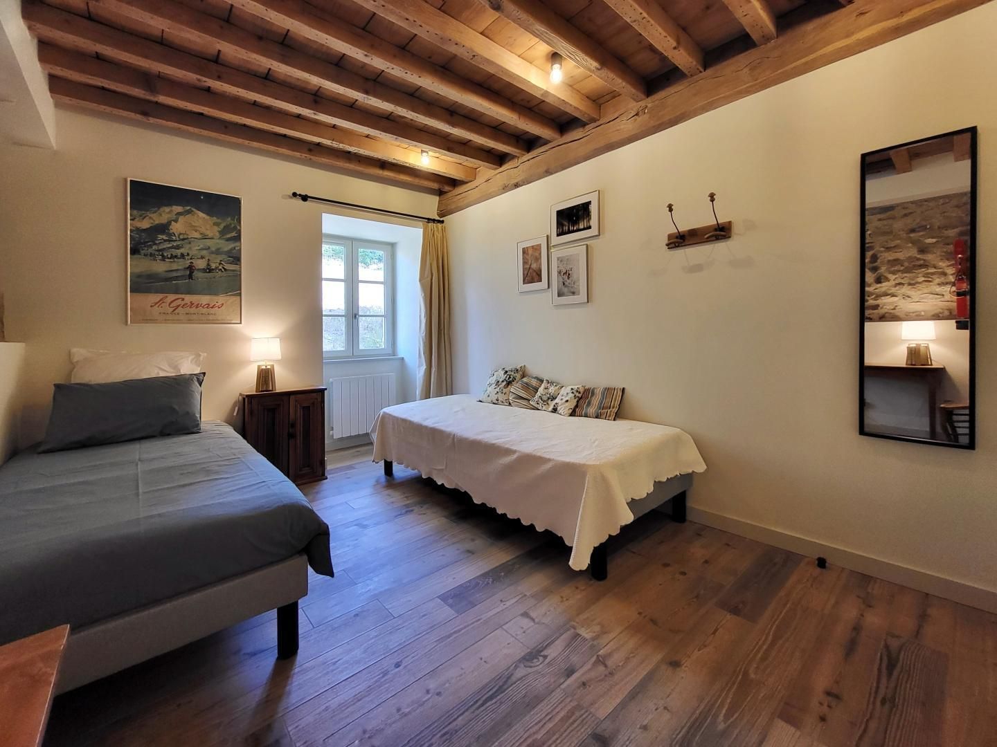 Un dormitori amb dos llits, terres de fusta, una finestra amb cortines i obra penjada a la paret.
