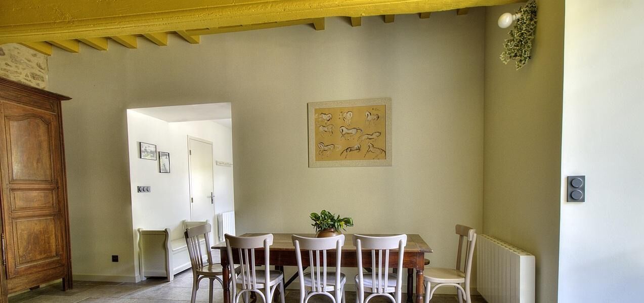 A dining area with a wooden table and white chairs, a potted plant, and a framed artwork on the wall.