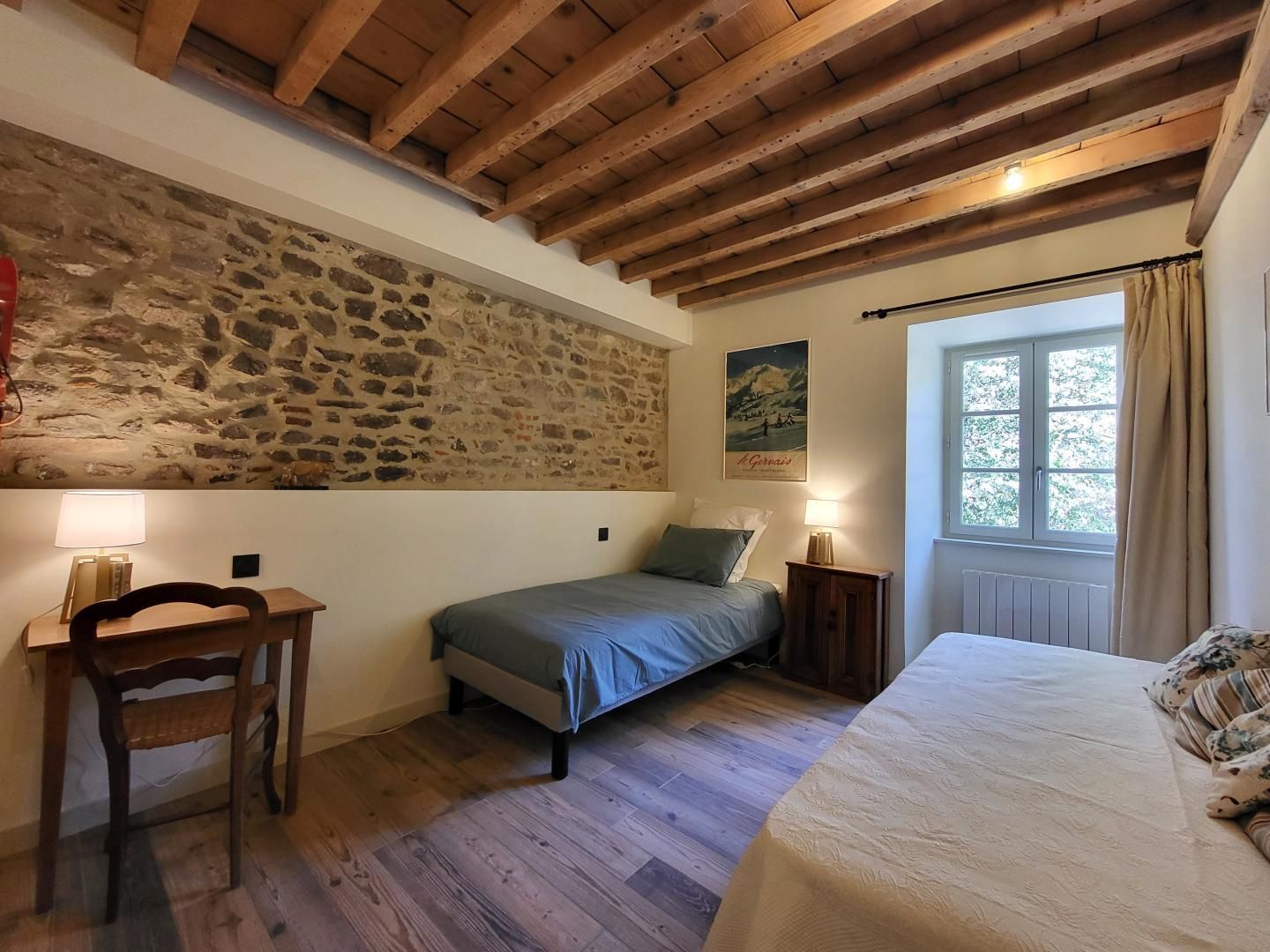 A bedroom with a stone wall, wooden beams, a bed, a desk, and a window with curtains.