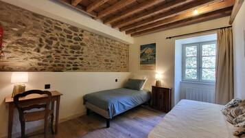 A bedroom with a stone wall, wooden beams, a bed, a desk, and a window with curtains.