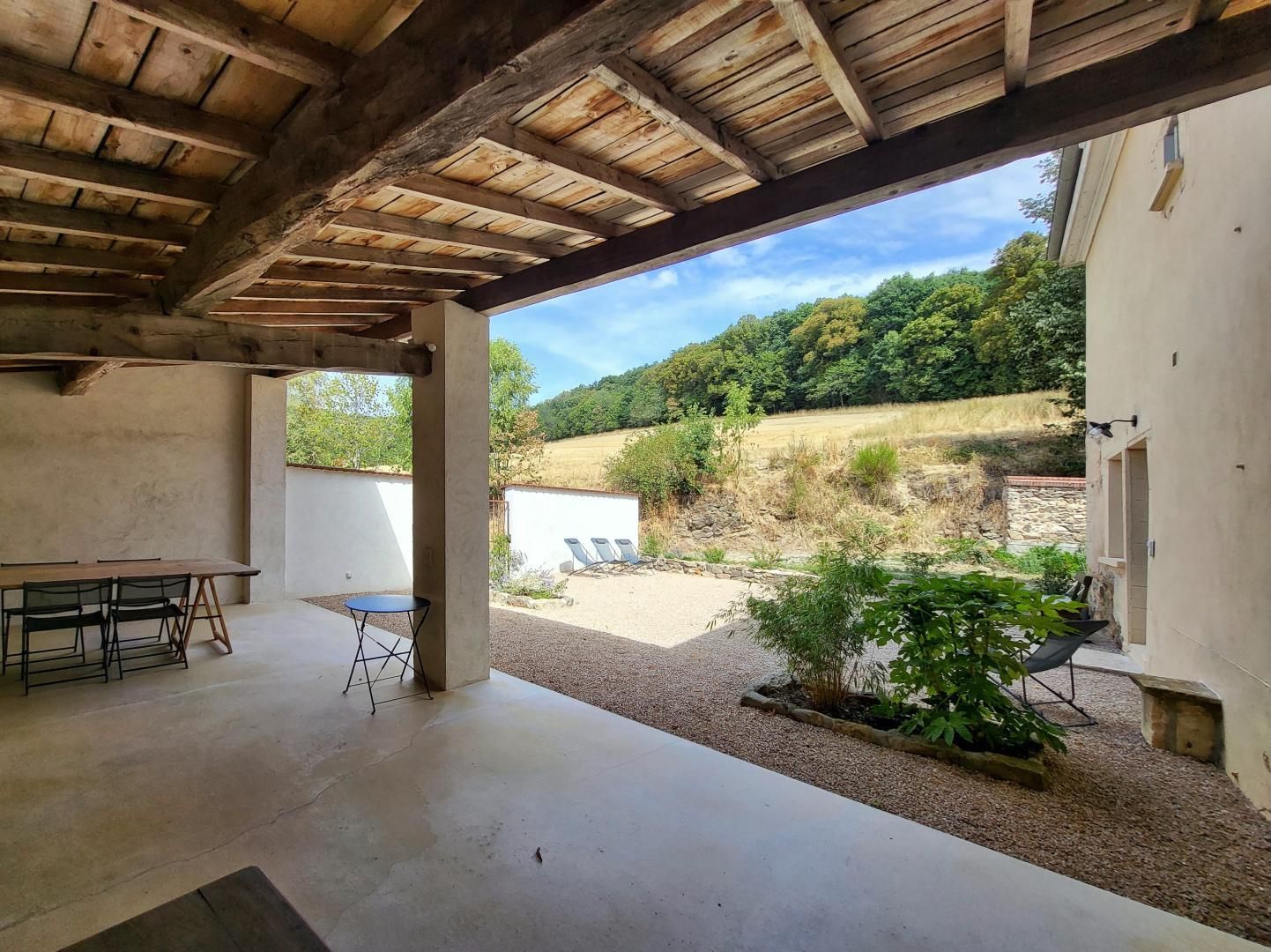 Een overdekt terras buiten, met een betonnen vloer, een houten tafel met stoelen, een klein krukje, en een uitzicht op een heuvel met bomen en een wit gebouw op de achtergrond.