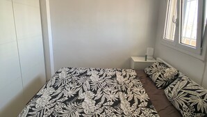 A small, sparsely furnished room with a bed, a bedside table, and a window. - Well-equipped apt, in the center of town, near beach and shops, overlooking natural park and sea. (Empuriabrava)