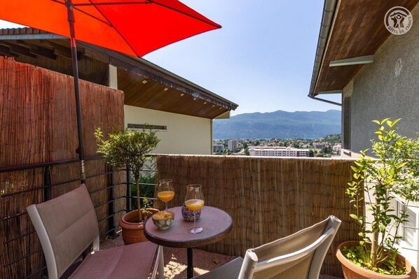 Une terrasse extérieure avec une table, des chaises et un parasol rouge. On y trouve des plantes en pot et, en arrière-plan, une vue sur une ville et des montagnes.