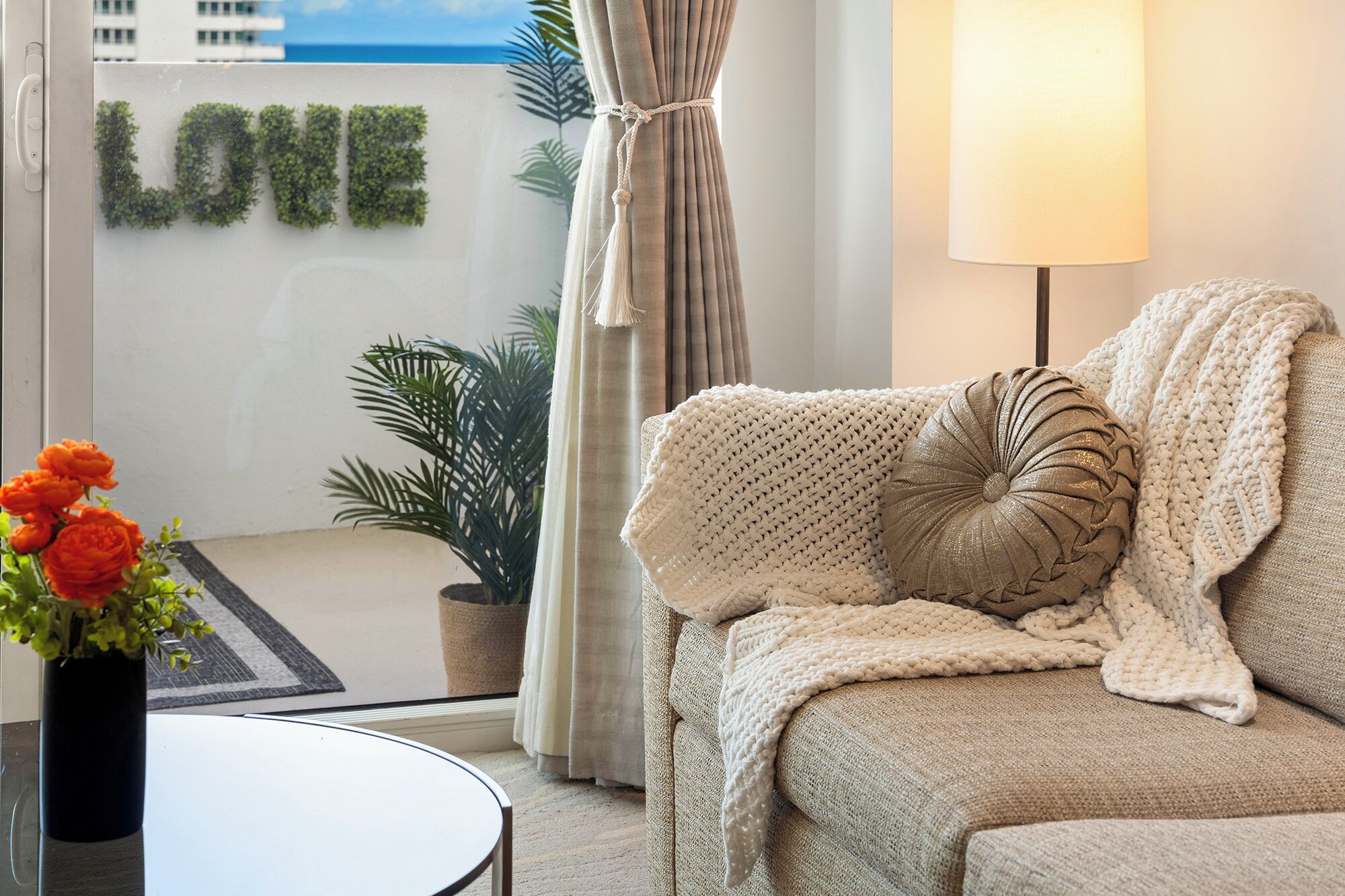 Una sala acogedora con un sofá color beige, una manta tejida y un cojín decorativo. Una planta en maceta y un florero con flores están en el suelo. Se ve una ventana con cortinas y vistas a edificios.