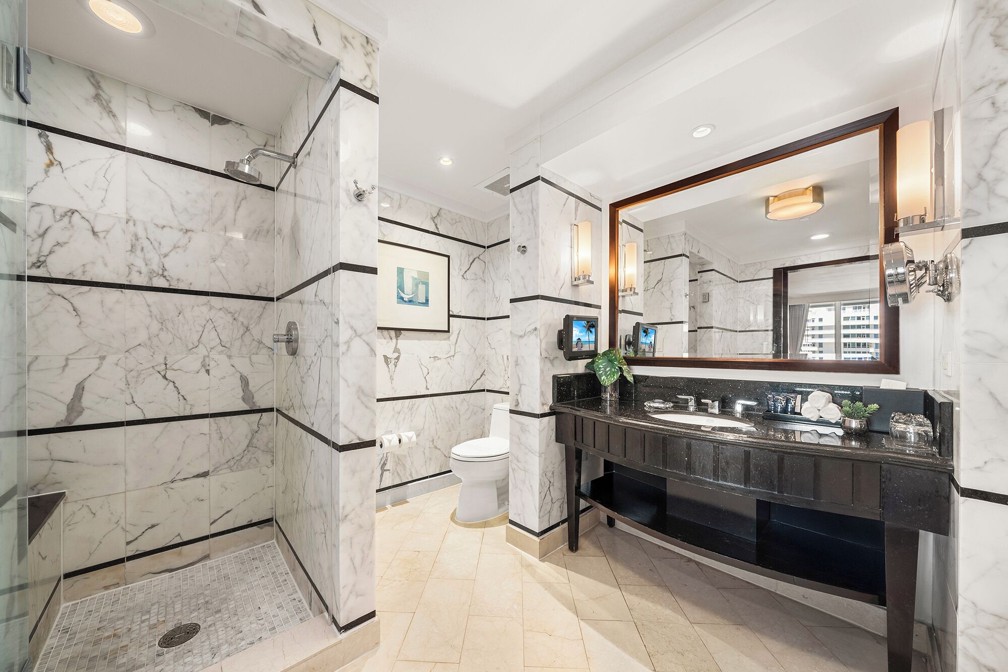 A modern bathroom with marble walls, a large mirror, a black vanity, and a toilet.
