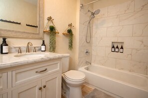 Ein Badezimmer mit einem weißen Waschbecken, einer Toilette, einer Badewanne und einer Dusche. Pflanzen stehen auf Regalen und Pflegeprodukte hängen an der Wand.