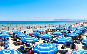 Ein Strand mit vielen blauen Sonnenschirmen und Liegestühlen.