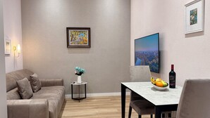 A modern living room with a sofa, a small side table with a vase, a dining table with chairs, and a large framed picture on the wall.