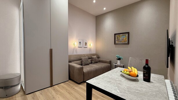 A modern living room with a sofa, a dining table with fruit and a bottle, a small side table with a vase, and a painting on the wall.