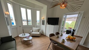 A modern living room with a sofa, a dining table with chairs, a TV mounted on the wall, and a kitchen area with a fruit bowl. - Stresa Golf & Relax Romantik Suite (Gignese)