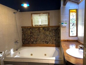 Un baño con pared de piedra, una bañera, un tocador de madera y una ventana con cortina de encaje.