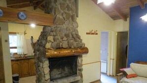 Una chimenea de piedra en una sala de estar con vigas de madera, un sofá y una cocina al fondo.