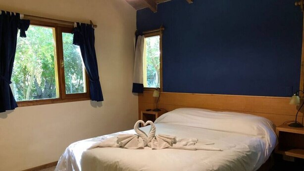 Un dormitorio con una cama, dos ventanas, una mesita de noche y una lámpara.