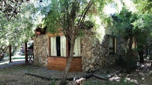 Una cabaña rústica de piedra con puerta y ventanas de madera, rodeada de árboles y un camino de grava.