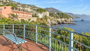 A coastal town with buildings on a hillside, a rocky shoreline, and a bench in the foreground.
