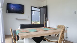 Una zona de menjador amb una taula de fusta, cadires blanques, un televisor fixat a la paret i una finestra amb vista a l'exterior.
