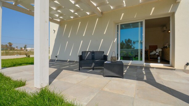 Ein überdachter Patio-Bereich mit Gartenmöbeln, eine Glastür, die zu einem Innenraum führt, und ein Blick auf die Umgebung.