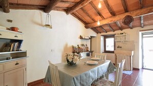 Un coin repas rustique avec un plafond en bois, une table dressée pour deux personnes et un espace cuisine avec une étagère portant l’inscription « THÉ ».
