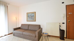 A living room with a sofa, a small table with a remote control, a radiator, and a door with a sign. - Appartamento Signoretti (Pieve di Soligo)