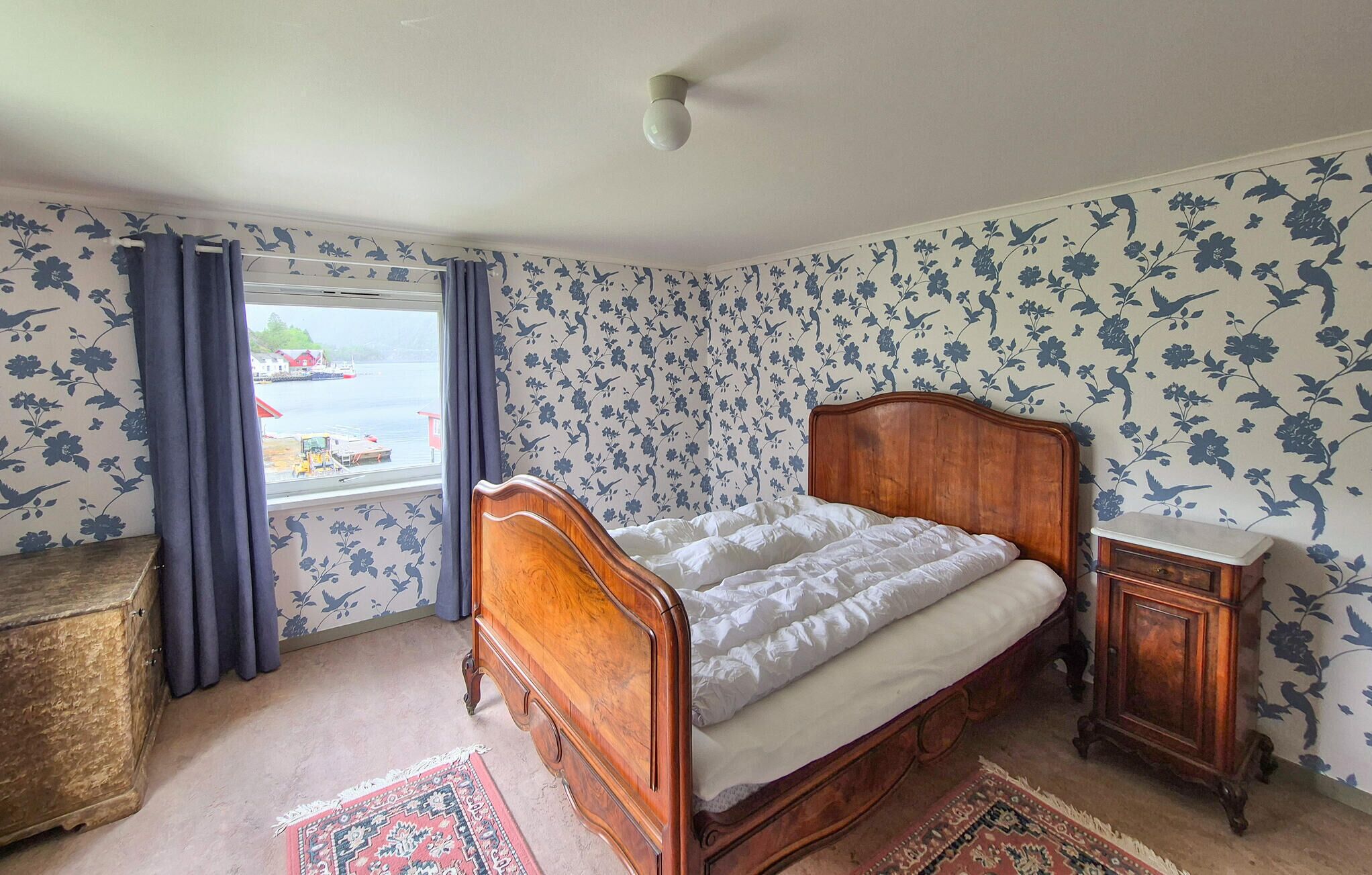 Une chambre avec un lit en bois, une fenêtre avec des rideaux, un papier peint à motifs floraux et une vue sur un quai.