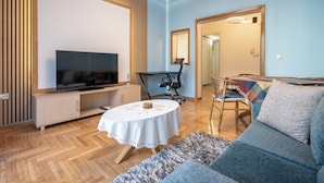 Un moderno salón con un televisor de pantalla plana, paneles de madera, una mesa redonda con mantel blanco, un sofá azul y una alfombra mullida.