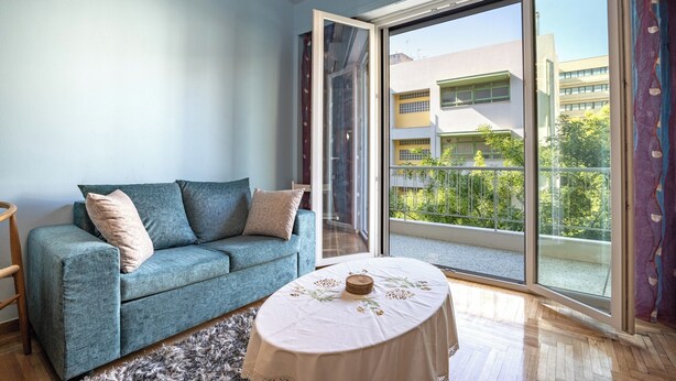 Una habitación con un sofá azul, una mesa redonda con mantel blanco y grandes puertas corredizas de vidrio que dan a un balcón con vistas a edificios y zonas verdes.