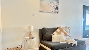 Ett modernt vardagsrum med en grå soffa, ett litet sidobord med en lampa, en korg av rotting och en inramad tavla på väggen.